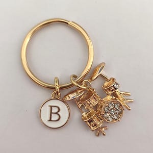 May include: Gold-tone keychain with a small drum set charm and a white circle charm with the letter "B" in gold.