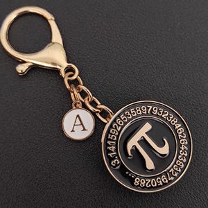 May include: Gold-tone keychain with a black and gold Pi symbol charm. The charm features the Pi symbol and the first few digits of Pi. An additional charm with the letter "A" hangs from the chain.