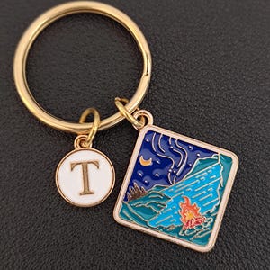 May include: Gold keychain with a blue and green enamel charm depicting a night scene with a campfire, moon, and stars. The charm is attached to a gold ring with a smaller round charm that says "T" in white letters.
