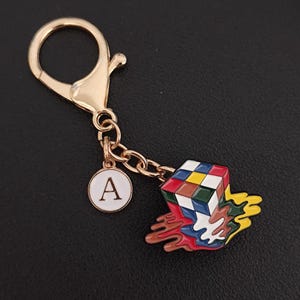 May include: Gold-tone keychain with a Rubik's Cube charm in a melting design. The charm features a colorful cube with a white border, and a white circular charm with the letter "A".