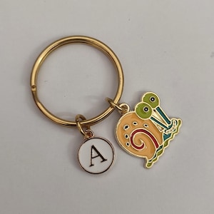 May include: Gold-tone keychain featuring a cartoon snail charm and a circular charm with the letter "A". The snail charm is orange, yellow, green, and red. The keychain ring is gold.