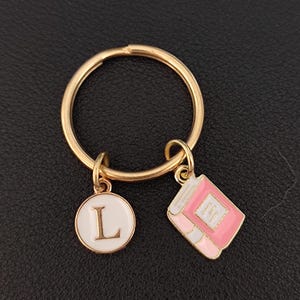 May include: Gold key ring with a pink and white book charm and a white circle charm with the letter L in gold.