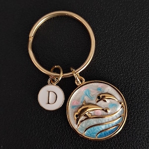 May include: Gold-tone keychain with a round charm featuring two gold dolphins leaping over blue and white waves. A smaller white charm with the letter "D" hangs nearby. The keychain is on a black surface.