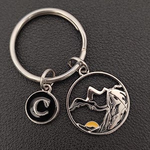 May include: A silver key ring with a round charm featuring a black and white mountain silhouette with a yellow sun. A smaller round charm with a black background and a white letter C is attached to the key ring.