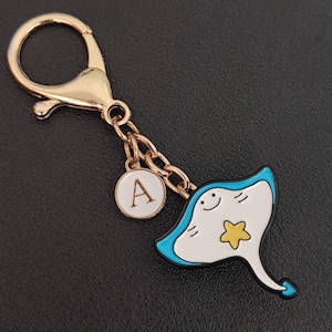 Può includere: Un portachiavi color oro con un ciondolo a forma di razza dei cartoni animati. La razza è bianca con un contorno blu e una stella gialla. Un piccolo ciondolo rotondo bianco con la lettera "A" pende dalla catena.