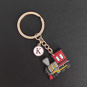 May include: A gold-colored keychain with a train-shaped charm. The train is red, gray, yellow, and black. A small, round charm with the letter "K" is also attached. The keychain is on a black surface.