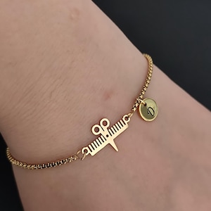 May include: Gold-colored bracelet featuring a charm with a barber's comb and scissors design, along with a small round charm with the letter "D". The bracelet has a delicate chain link design.