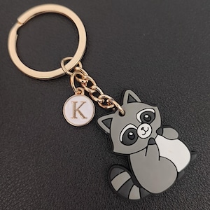 Könnte beinhalten: Ein goldfarbener Schlüsselanhänger mit einem Cartoon-Waschbär-Anhänger. Der Waschbär ist grau, weiß und schwarz, mit großen Augen und einem freundlichen Gesichtsausdruck. Ein kleiner, runder Anhänger mit dem Buchstaben "K" hängt neben dem Waschbären.