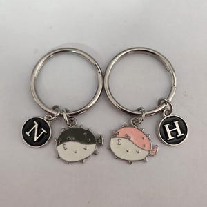 Op de afbeelding: Twee zilveren sleutelhangers met zwarte en witte kogelvis-charms. De charms hebben zwarte en roze accenten. De sleutelhangers hebben ook zwarte ronde charms met de letters "N" en "H" in wit.
