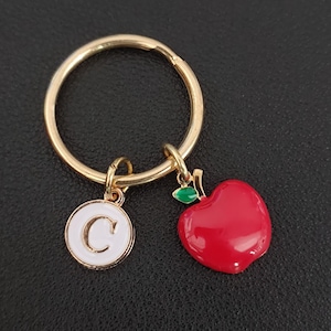 Peut inclure: Porte-clés doré avec un charm en forme de pomme rouge et un charm circulaire blanc avec la lettre "C". Le charm pomme a une feuille verte. Le porte-clés est sur une surface noire.