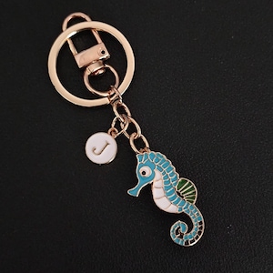 May include: A gold-tone metal keyring with a seahorse charm in blue, white, and green enamel. It includes a small, round charm with the letter "J" on a white background. The seahorse is a decorative element.