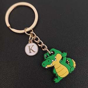 May include: A gold-colored keychain with a green and yellow alligator charm. The keychain includes a small, round charm with the letter "K" on it. The alligator charm is detailed with black and white accents.