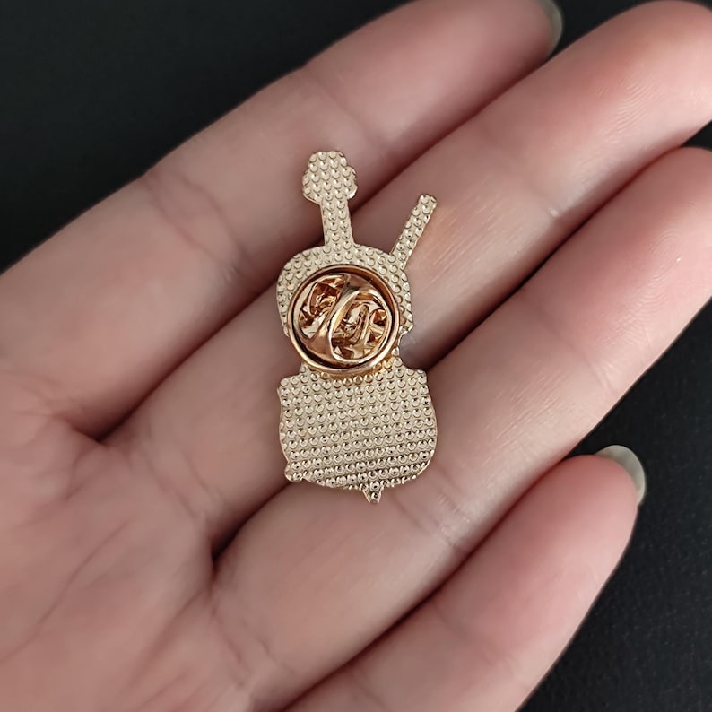 May include: A gold-colored metal pin in the shape of a violin. The pin has a textured surface and a butterfly clasp on the back. The violin design includes a scroll, neck, body, and a bow.