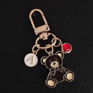 May include: Rose gold keychain with a teddy bear charm, a red gem, and a circular charm with the letter "A". The teddy bear is brown with gold accents and a bow tie. The keychain has a lobster clasp.