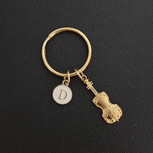 Puede incluir: Llavero dorado con un dije de violín y un dije circular blanco con la letra "D". El llavero se encuentra sobre una superficie negra.