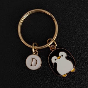 Könnte beinhalten: Ein goldfarbener Schlüsselanhänger mit einem runden Ring und zwei Anhängern. Ein Anhänger ist ein weißer Kreis mit dem Buchstaben "D". Der andere Anhänger ist ein schwarz-weißer Pinguin mit gelben Füßen.