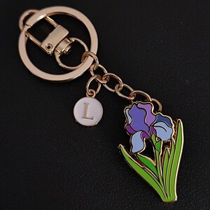 May include: Gold-coloured keyring featuring a purple and blue iris flower charm and a white circular charm with the letter "L". The flower has green leaves. The keyring has a lobster clasp and a split ring.