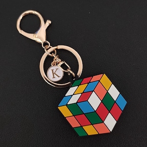 Op de afbeelding: Een goudkleurige sleutelhanger met een Rubik's Cube-bedel. De kubus heeft een kleurrijk patroon van rode, blauwe, groene, gele, oranje en witte vierkanten. Een kleine ronde bedel met de letter "K" is ook bevestigd.