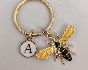 May include: Gold key ring with a yellow and black bee charm and a round charm with the letter A on a white background.