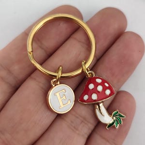 May include: Gold key chain with a red and white mushroom charm and a gold charm with the letter E on a white background.