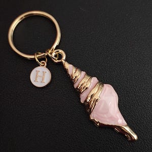 May include: A gold-tone keyring with a pink seashell pendant and gold detailing. It includes a small, circular charm with the letter "H". This keyring is suitable for keys or as a decorative item.