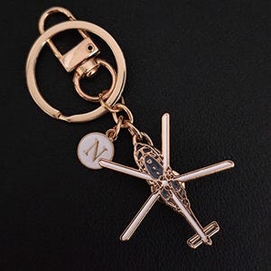 May include: Gold-coloured metal keyring featuring a helicopter charm with white and navy blue accents. Includes a round charm with the letter "N" in white. The keyring has a clasp and a key ring.