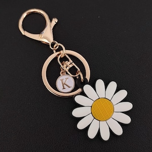 Op de afbeelding: Een goudkleurige sleutelhanger met een madeliefjesbedel en een ronde bedel met de letter "K". Het madeliefje heeft witte bloemblaadjes, een gele kern en een zwarte omtrek. De sleutelhanger bevat een sluiting en een sleutelring.