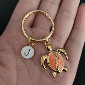 May include: A gold-toned metal keyring featuring a circular ring, a small white circular charm with the letter J in gold, and a sea turtle charm with a textured, coral-coloured shell inlay. The metal components have a polished, reflective finish.