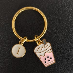May include: A gold-coloured keyring with a circular ring. The keyring features two charms: a white circle with the letter "I" and a pink and white boba tea cup. The boba tea cup has black boba pearls.