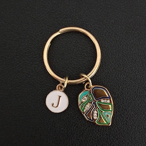 May include: Gold-tone keychain with a round ring and two charms. One charm is a white circle with the letter "J", and the other is a leaf-shaped charm with brown, green, and blue enamel and small crystals. The background is black.