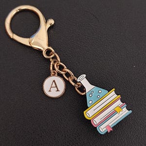 May include: Gold-tone keychain with a charm featuring a stack of books, a flask with blue liquid, and a circular charm with the letter "A". The keychain has a lobster clasp for easy attachment.