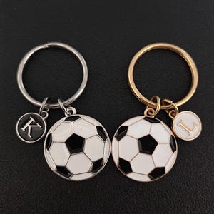 Può includere: Due portachiavi d'argento e d'oro con ciondoli a forma di pallone da calcio e ciondoli con lettere. Il portachiavi d'argento ha un ciondolo con la lettera K nera e il portachiavi d'oro ha un ciondolo con la lettera L nera.