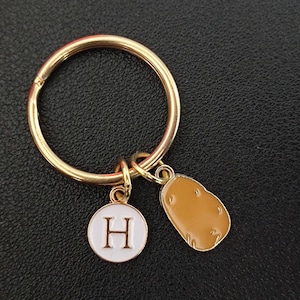 May include: Gold-colored keychain with a round ring and two charms. One charm is a white circle with a capital "H" in the center. The other charm is a brown potato shape.