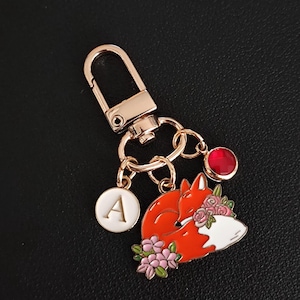 May include: A gold-colored keychain featuring a sleeping fox design with orange, white, and pink enamel details. The keychain includes a round charm with the letter "A" and a red gem. The fox is adorned with pink flowers.