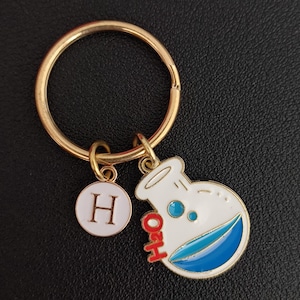 May include: Gold-coloured keychain with two charms. One charm is a white circle with the letter "H". The other charm is a white flask with blue liquid and the letters "H2O" in red. The keychain is on a black surface.