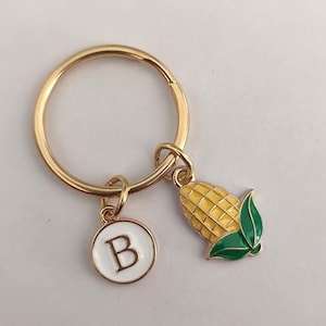 May include: Gold key ring with a yellow corn charm and a white circle charm with the letter B in black.