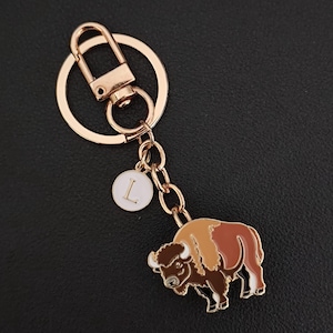 May include: A gold-colored metal keychain featuring a bison charm with brown, tan, and white enamel details. The keychain includes a round charm with the letter "L" and a clasp.