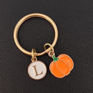 May include: Gold key chain with a white circle charm featuring the letter L and a small orange pumpkin charm.