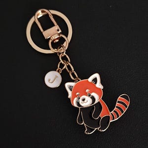 Peut inclure: Un porte-clés représentant un panda roux avec un fermoir et une chaîne dorés. Le panda roux a un visage et un corps noirs et blancs, avec une queue rayée. Un petit charme blanc avec la lettre "J" est suspendu à la chaîne.