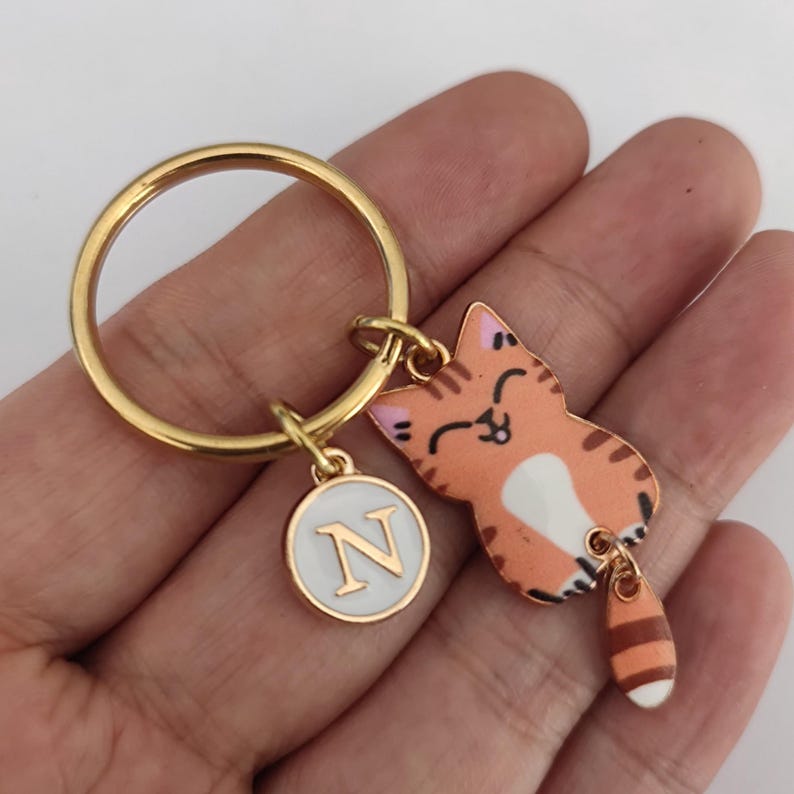 May include: Gold keychain with a round charm featuring the letter N and a cartoon orange and white cat charm with a dangling tail.