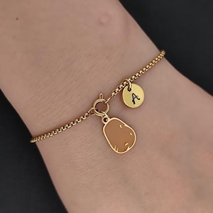 Könnte beinhalten: Ein goldfarbenes Kettenarmband mit einem runden Anhänger, der mit dem Buchstaben A graviert ist, und einem weiteren Anhänger in Form einer braunen Kartoffel. Das Armband wird mit einem runden Federringverschluss geschlossen und glänzt metallisch.