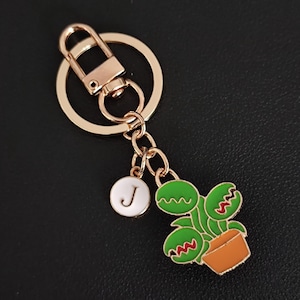May include: Gold-tone keychain with a lobster clasp, a round charm with the letter "J", and a green and orange enamel plant charm. The plant charm has red accents and is in a brown pot.