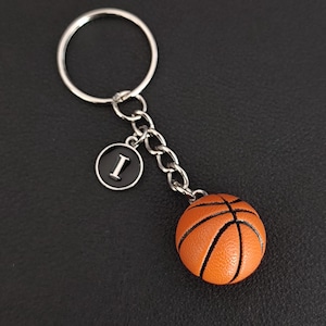 Könnte beinhalten: Ein Basketball-Schlüsselanhänger mit einem kleinen orangefarbenen Basketball mit schwarzen Linien, einer silbernen Kette und einem runden Anhänger mit dem Buchstaben "I" auf schwarzem Hintergrund. Der Schlüsselanhänger hat einen silbernen Schlüsselring.