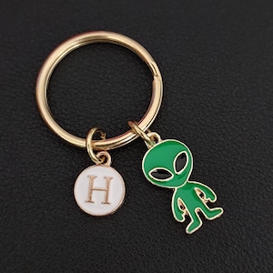 May include: Gold-tone keychain with a green alien charm and a white circular charm with the letter "H". The alien charm has black eyes and a gold outline. The keychain is on a black surface.