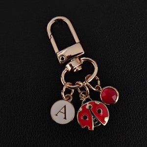 Może przedstawiać: Złoty brelok do kluczy z karabińczykiem, z białą zawieszką z literą "A", czerwoną zawieszką biedronki i czerwoną zawieszką z kamieniem szlachetnym. Biedronka ma czarne kropki i czerwone skrzydła. Brelok leży na czarnej powierzchni.