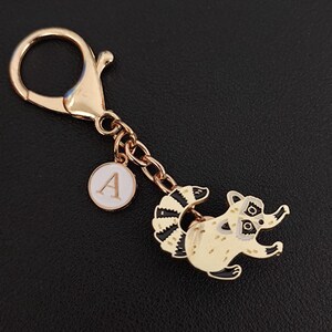 May include: A gold-coloured keychain featuring a raccoon charm. The raccoon is cream-coloured with black and white details. A small, round charm with the letter "A" is attached to the chain.