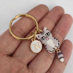 May include: Gold keychain with a cartoon cat charm and a round charm with the letter N. The cat charm is gray and white with a black and white striped tail.