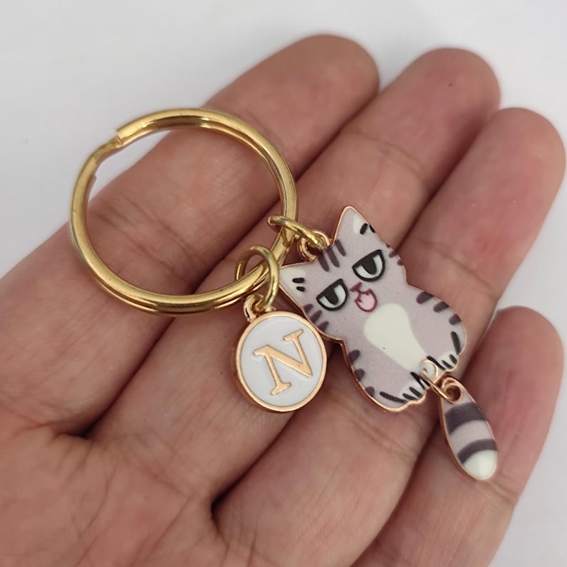 May include: Gold keychain with a cartoon cat charm and a round charm with the letter N. The cat charm is gray and white with a black and white striped tail.