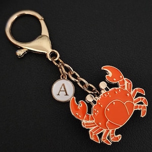 Könnte beinhalten: Ein goldfarbener Schlüsselanhänger mit Karabinerverschluss, Kette und einem Anhänger mit einem orangefarbenen Krebsdesign. Ein kleiner weißer runder Anhänger mit dem Buchstaben "A" ist ebenfalls angebracht. Der Krebsanhänger hat goldfarbene Umrisse.