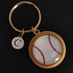 Könnte beinhalten: Ein goldfarbener Schlüsselanhänger mit einem Baseball-Anhänger. Der Anhänger zeigt einen weißen Baseball mit roten Nähten, eingefasst in einen goldfarbenen Rahmen. Ein kleinerer, weißer Anhänger mit dem Buchstaben "C" hängt daneben.
